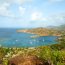 Aerial view of a scenic harbor in Antigua with sailboats anchored in turquoise water, surrounded by green rolling hills, beaches, and coastal homes