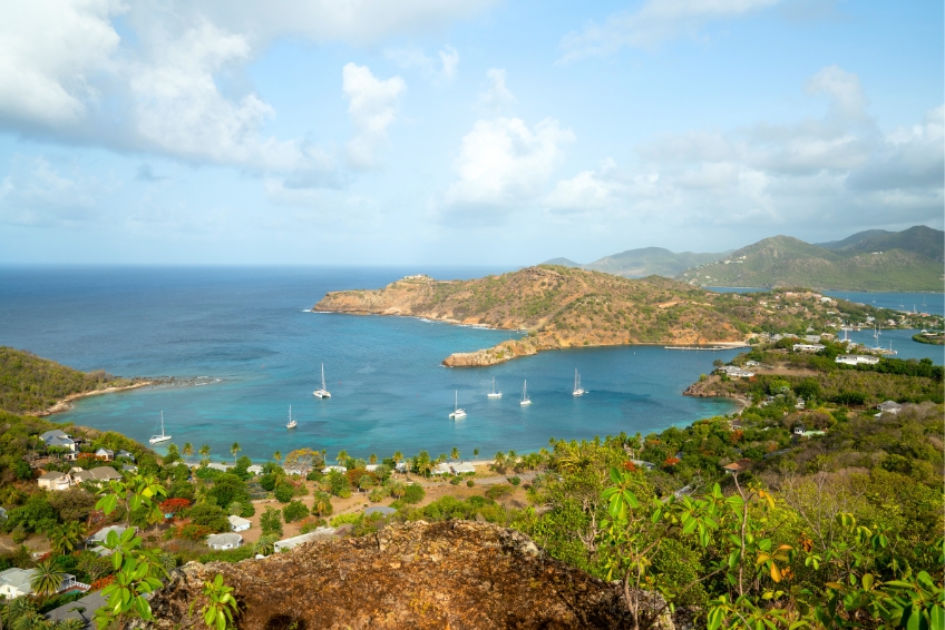 Aerial view of a scenic harbor in Antigua with sailboats anchored in turquoise water, surrounded by green rolling hills, beaches, and coastal homes