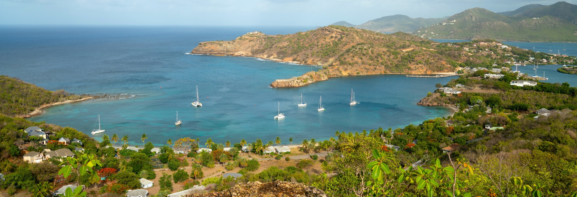 Aerial view of a scenic harbor in Antigua with sailboats anchored in turquoise water, surrounded by green rolling hills, beaches, and coastal homes