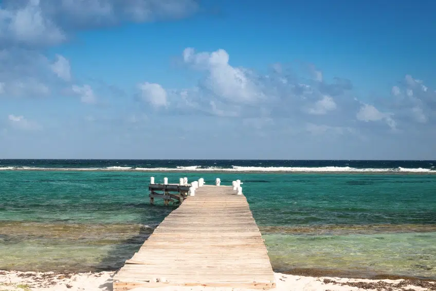 Rustic wooden pier with white posts extending into turquoise Caribbean water in the Cayman Islands with waves breaking on the reef in the distance