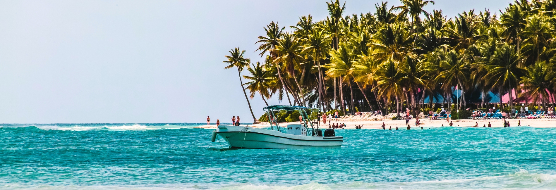 Tropical beach scene in Punta Cana, Dominican Republic with a boat in turquoise water, palm trees lining the shore, and beachgoers enjoying the white sand