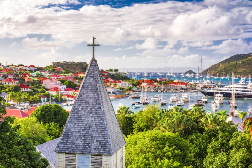 Panoramic view of Gustavia harbor in St. Barts with a church steeple in the foreground, colorful buildings on hillsides, and yachts anchored in the turquoise bay