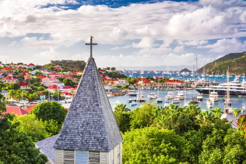 Panoramic view of Gustavia harbor in St. Barts with a church steeple in the foreground, colorful buildings on hillsides, and yachts anchored in the turquoise bay