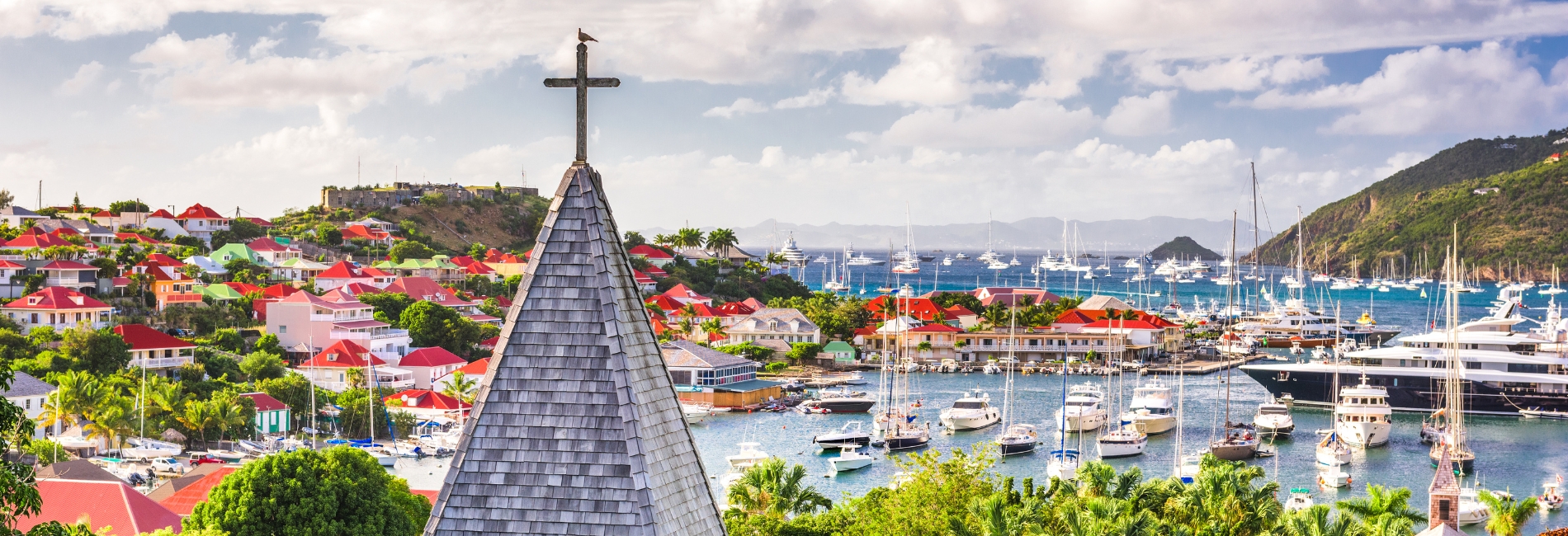 St. Barts Gustavia Harbor with Church Steeple and Colorful Red Roofed Buildings Panoramic view of Gustavia harbor in St. Barts with a church steeple in the foreground, colorful buildings on hillsides, and yachts anchored in the turquoise bay