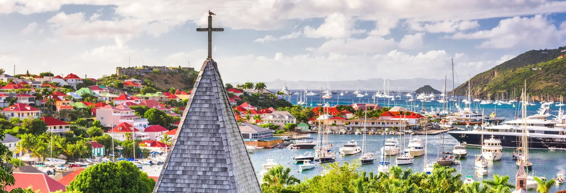 Panoramic view of Gustavia harbor in St. Barts with a church steeple in the foreground, colorful buildings on hillsides, and yachts anchored in the turquoise bay