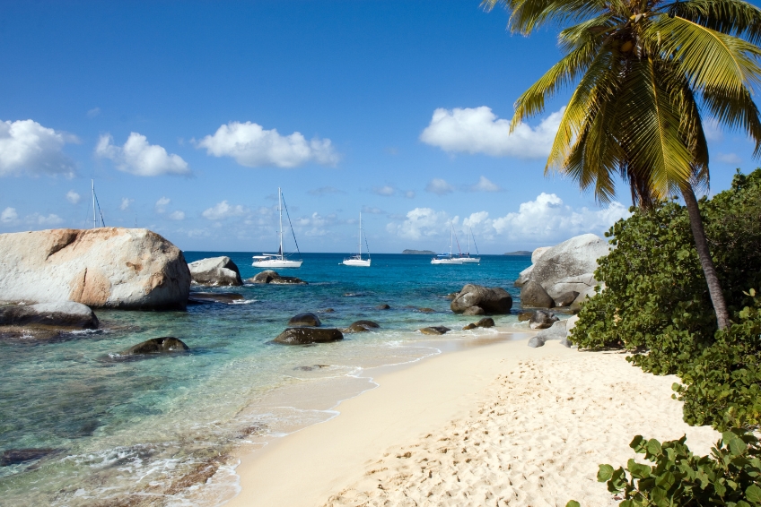 Scenic beach in the U.S. Virgin Islands with large granite boulders, clear turquoise water, anchored sailboats, and palm trees