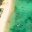 Aerial view of a pristine white sand beach in Bimini with rows of beach umbrellas, palm trees, and boats anchored in crystal clear turquoise water