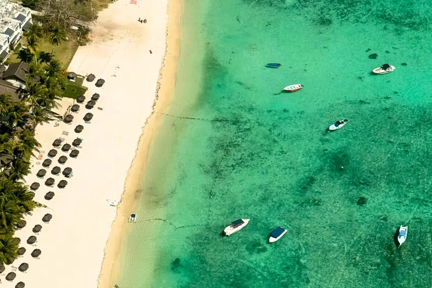 Aerial view of a pristine white sand beach in Bimini with rows of beach umbrellas, palm trees, and boats anchored in crystal clear turquoise water