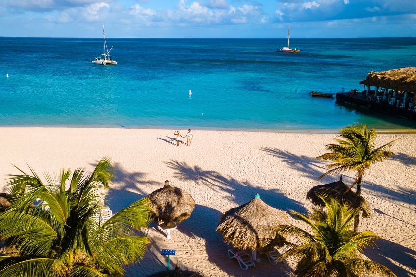 Palm Beach, Aruba Romantic Beach Getaway with Turquoise Water Couple holding hands on beautiful Palm Beach in Aruba viewed from above with white powdery sand, crystal clear turquoise water, palm tree shadows, beach umbrellas, and thatched waterfront bar
