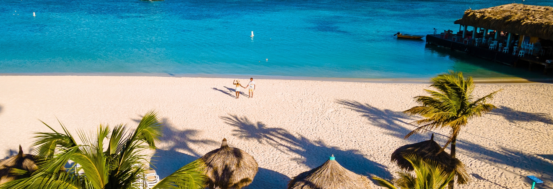 Best Time to Visit Aruba Palm Beach with White Sand and Turquoise Water Couple walking on pristine white sand at Palm Beach in Aruba with turquoise Caribbean water, palm trees casting shadows, thatched palapas, and overwater restaurant