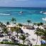 Aerial view of Hotel Riu Palace Antillas beach in Aruba with white sand, palm trees, thatched palapas, beachgoers, catamarans, and turquoise Caribbean water