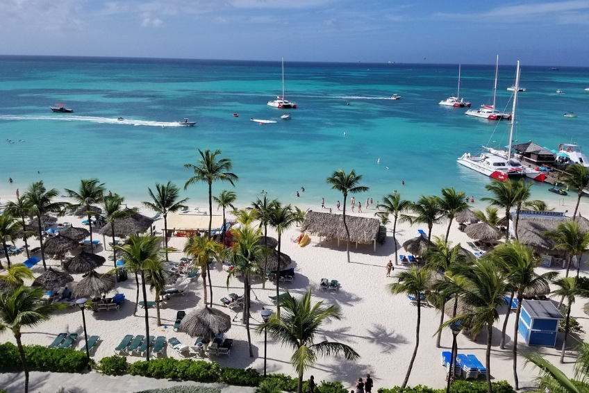 Aerial view of Hotel Riu Palace Antillas beach in Aruba with white sand, palm trees, thatched palapas, beachgoers, catamarans, and turquoise Caribbean water