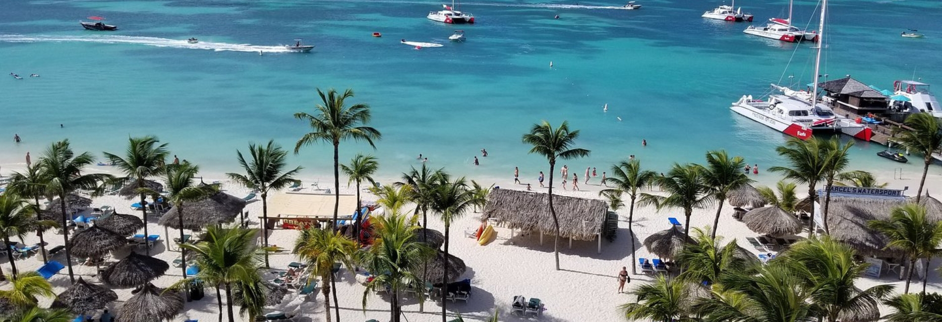 Hotel Riu Palace Antillas Beach Resort, Aruba with Water Activities Bustling beach at Hotel Riu Palace Antillas resort in Aruba with white sand, palm trees, thatched palapas, catamarans anchored in turquoise water, and watersports activities