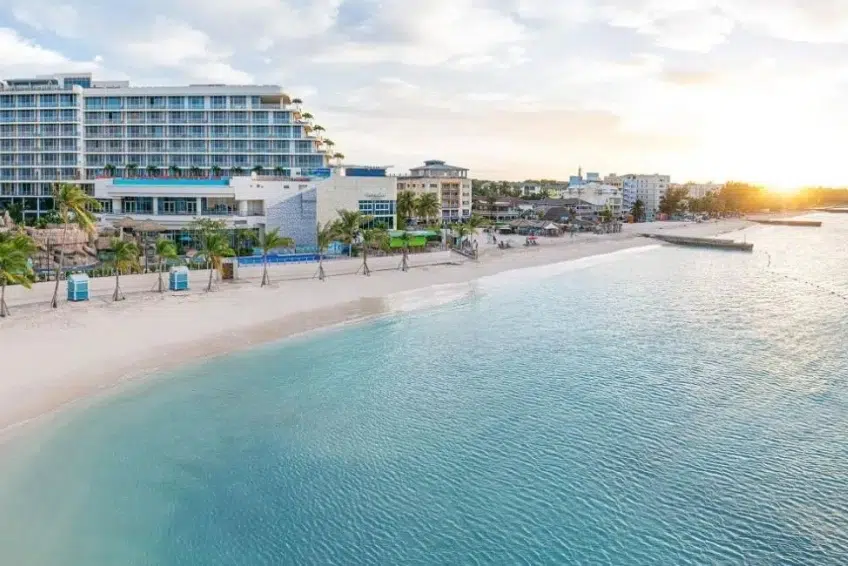 Panoramic view of luxury beachfront resort in the Bahamas featuring white sand beach, turquoise Caribbean water, palm trees, modern hotel buildings, and golden sunset light