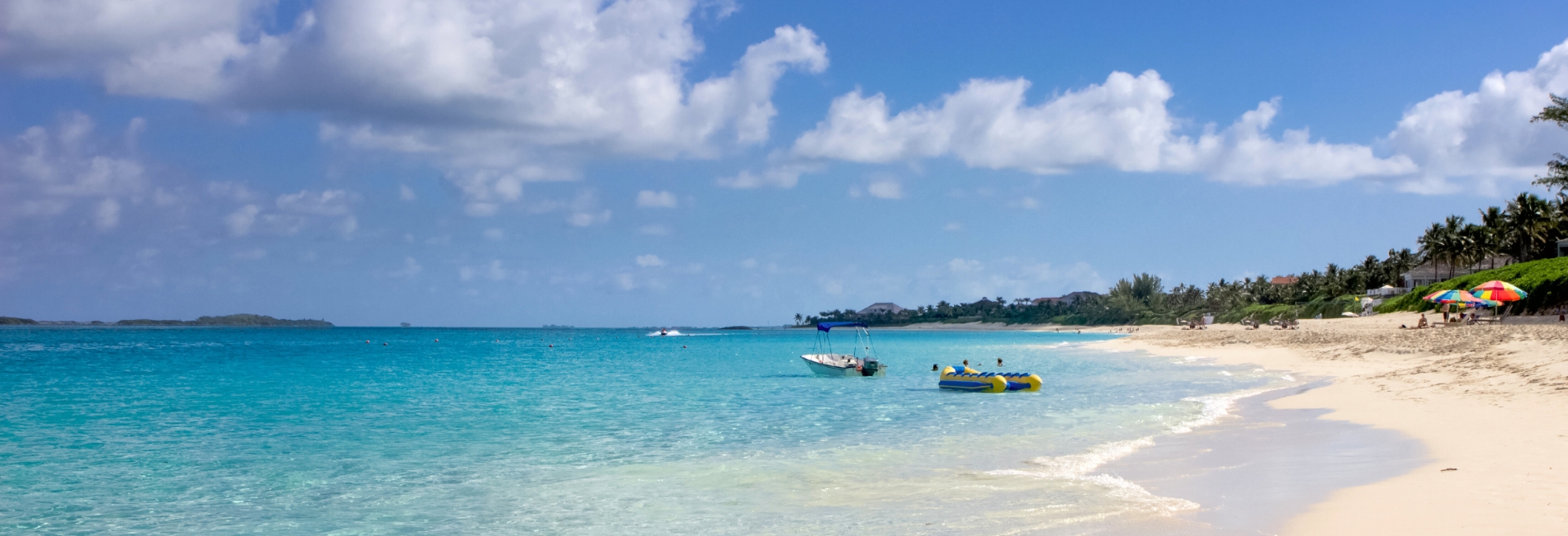 Paradise Beach in Nassau, Bahamas featuring crystal clear turquoise water, white sand beach, palm trees, colorful beach umbrellas, and boats