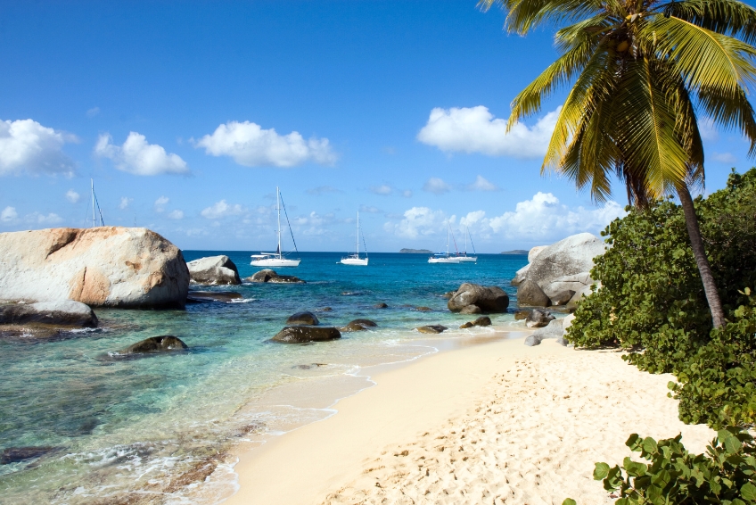 Scenic beach in the U.S. Virgin Islands with large granite boulders, clear turquoise water, anchored sailboats, and palm trees.