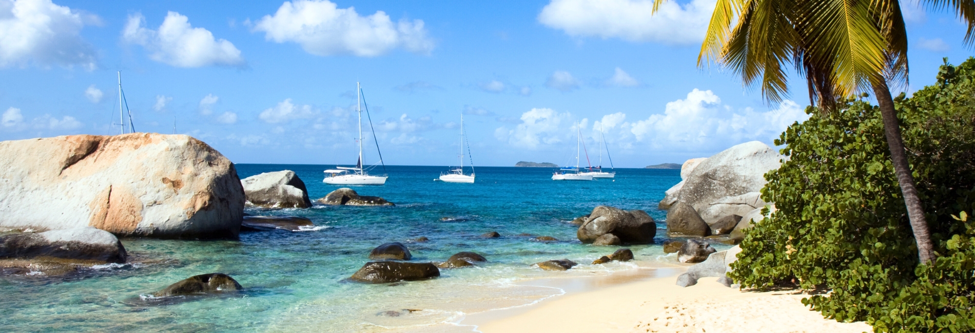 Scenic beach in the U.S. Virgin Islands with large granite boulders, clear turquoise water, anchored sailboats, and palm trees