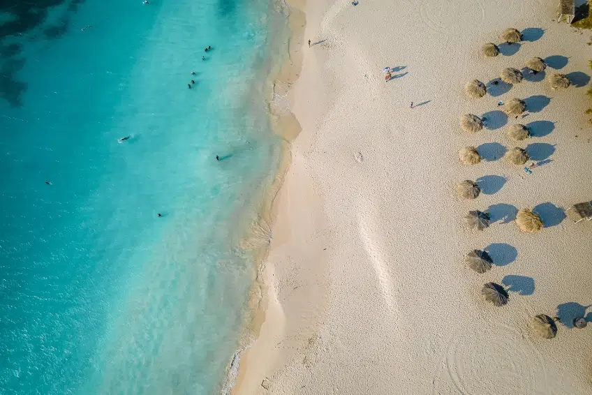 Stunning aerial view of Eagle Beach in Aruba showing white powdery sand, turquoise Caribbean water, beachgoers enjoying the water, and traditional thatched beach umbrellas