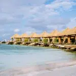 Luxury private cabanas at Renaissance Wind Creek Aruba Resort lined along the waterfront with thatched palapa roofs, outdoor loungers, water slide, and crystal clear turquoise water