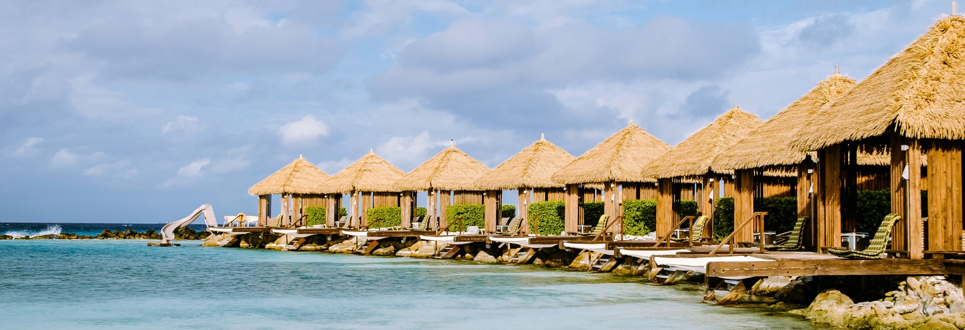 Row of private overwater cabanas at Renaissance Wind Creek Aruba Resort with thatched roofs, lounge chairs, water slide, and turquoise Caribbean water