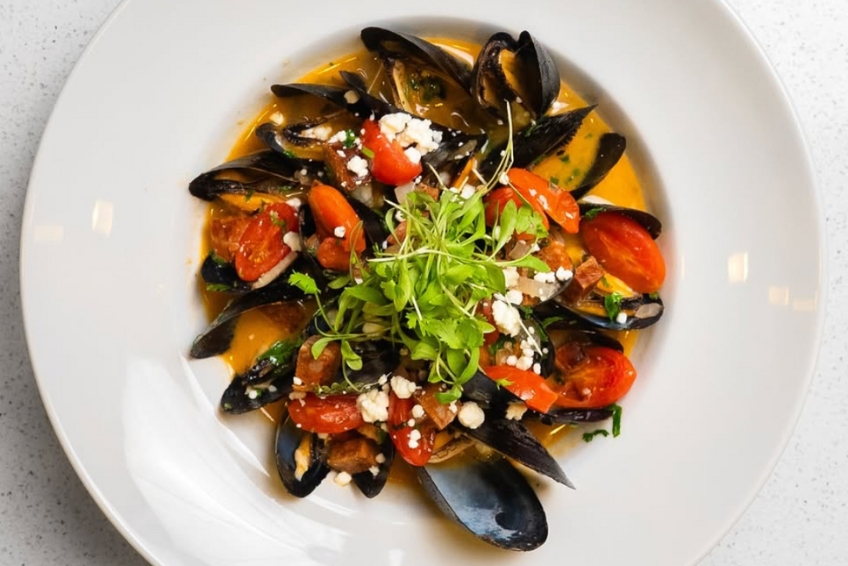 Gourmet mussels dish at Mesa Cocina restaurant in Puerto Rico with fresh mussels, roasted tomatoes, crumbled feta cheese, microgreens, and savory broth on white plate