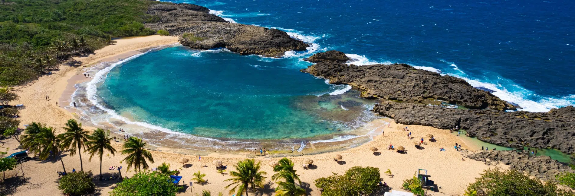 Aerial view of Mar Chiquita Beach in Puerto Rico featuring a protected turquoise cove surrounded by volcanic lava rocks, golden sand beach, palm trees, and beachgoers enjoying the natural pool