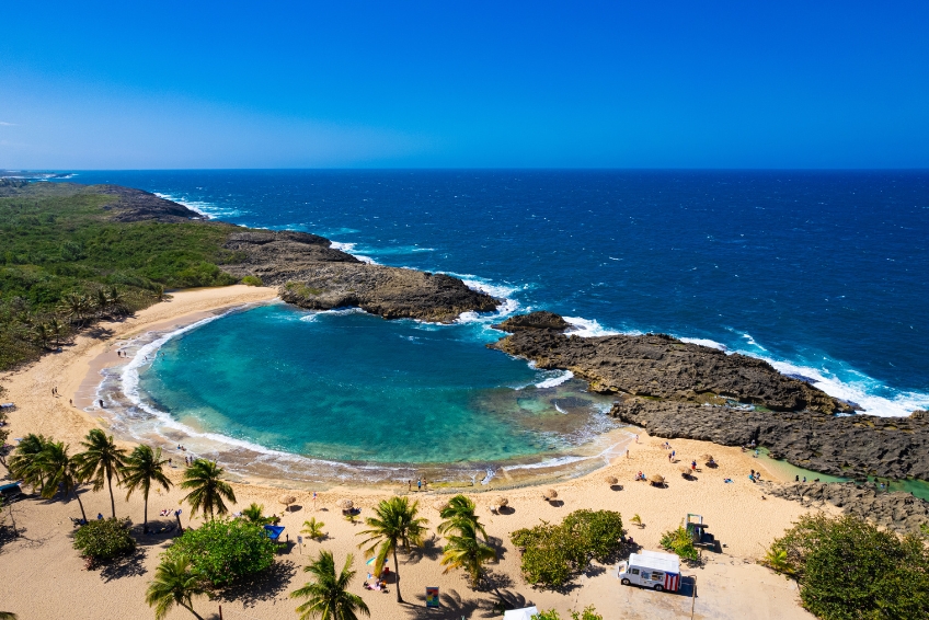 Mar Chiquita Beach, Puerto Rico Natural Pool Beach Aerial View Stunning Mar Chiquita Beach in Puerto Rico with a natural swimming pool formed by volcanic rock formations, golden sand, turquoise water, palm trees, and deep blue ocean waves