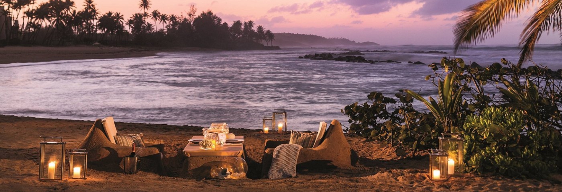 Romantic beachfront dining setup at Ritz-Carlton Dorado Beach in Puerto Rico with cushioned seating, lanterns, tropical foliage, purple sunset sky, palm trees, and ocean waves