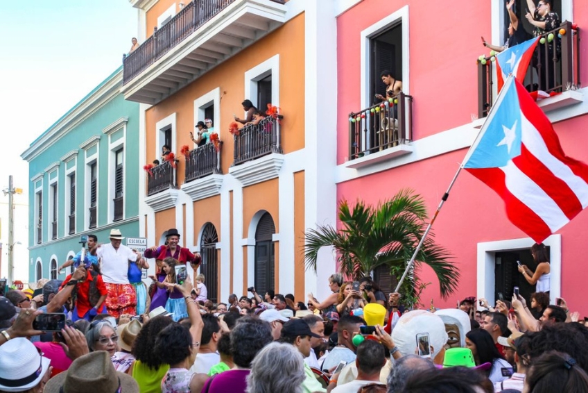 San Sebastián Street Festival in Old San Juan, Puerto Rico with crowds celebrating, traditional vejigante masks on stilts, Puerto Rican flag, colorful colonial buildings, and people on balconies