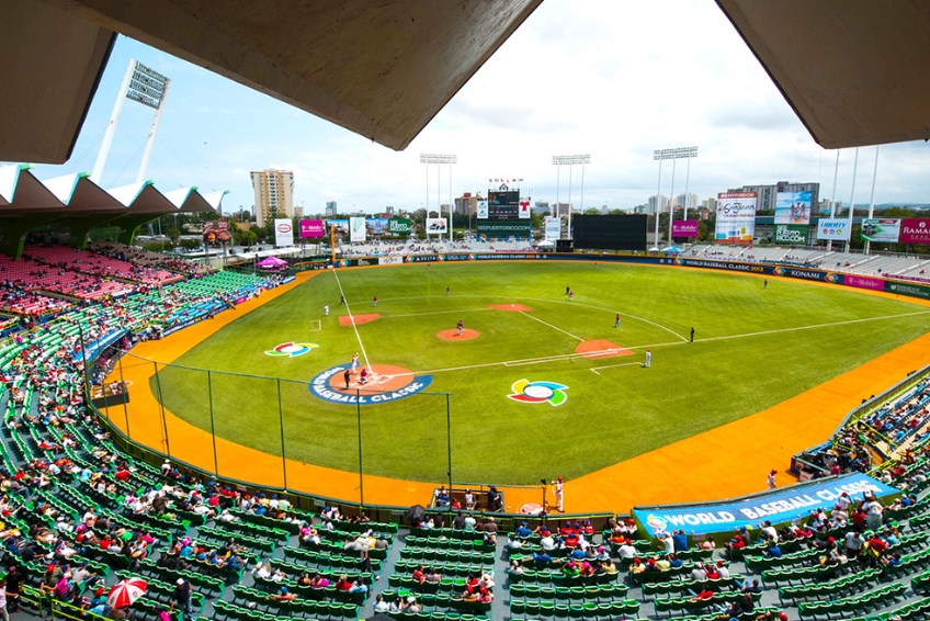 Hiram Bithorn Stadium in Puerto Rico during World Baseball Classic game with colorful seats, orange warning track, green outfield, fans in attendance, and San Juan cityscape in background
