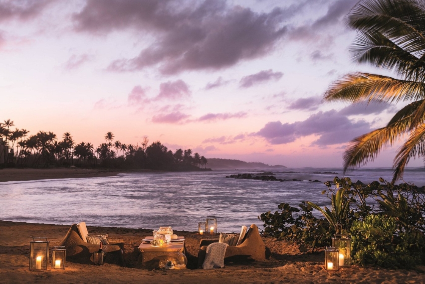 Luxury beachfront dining experience at Ritz-Carlton Dorado Beach in Puerto Rico featuring intimate seating area with pillows, candlelit lanterns, sunset views, palm trees silhouetted against purple sky, and ocean backdrop