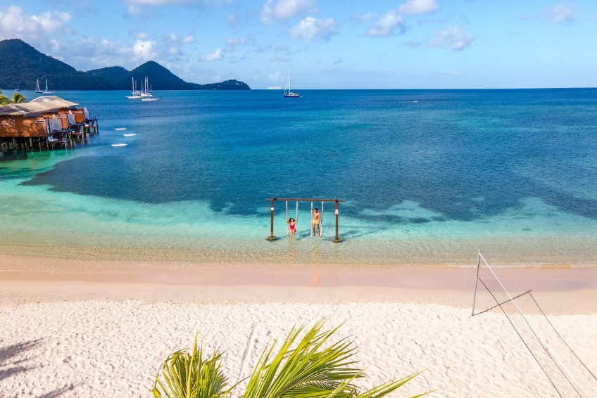 Picture-perfect St. Lucia beach featuring iconic wooden beach swings in crystal clear turquoise water, pristine white sand, overwater cabanas, anchored sailboats, and volleyball court