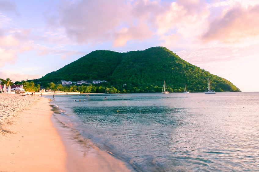 Scenic St. Lucia beach featuring white sandy shore, turquoise Caribbean water, anchored sailboats, dramatic green mountain backdrop, palm trees, and golden sunset light