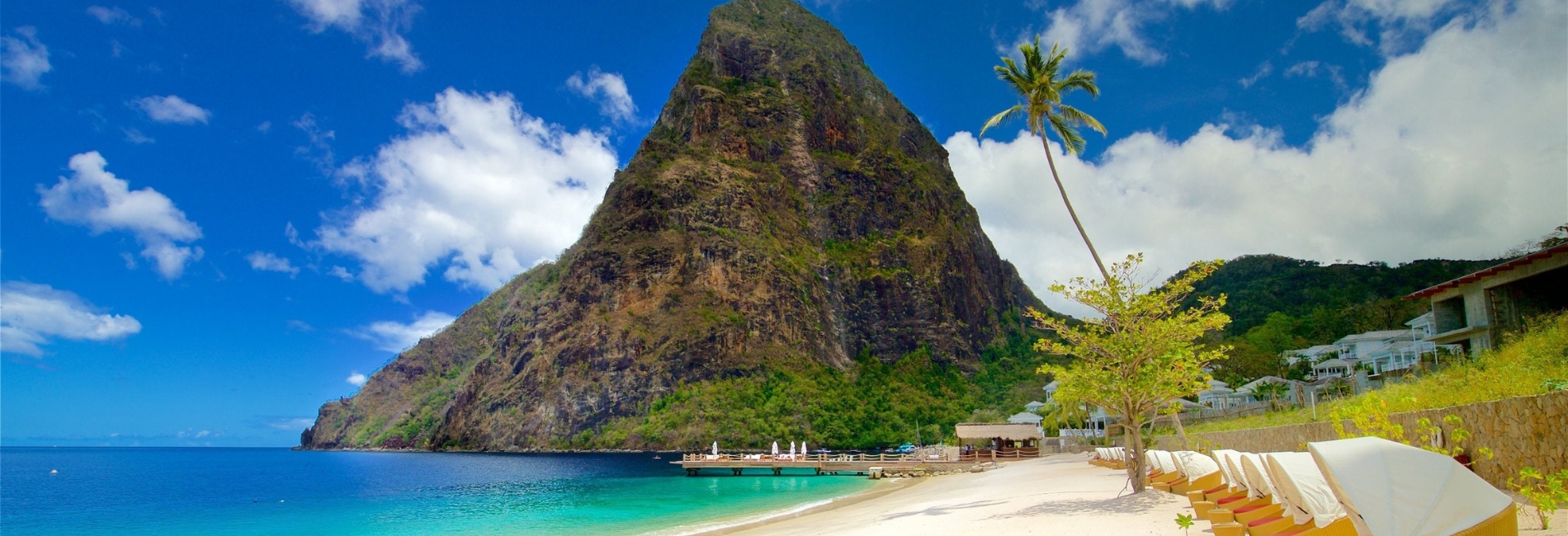 Stunning St. Lucia beach with dramatic Piton mountain, white sand beach, turquoise Caribbean water, palm tree, resort loungers, and blue sky with white clouds