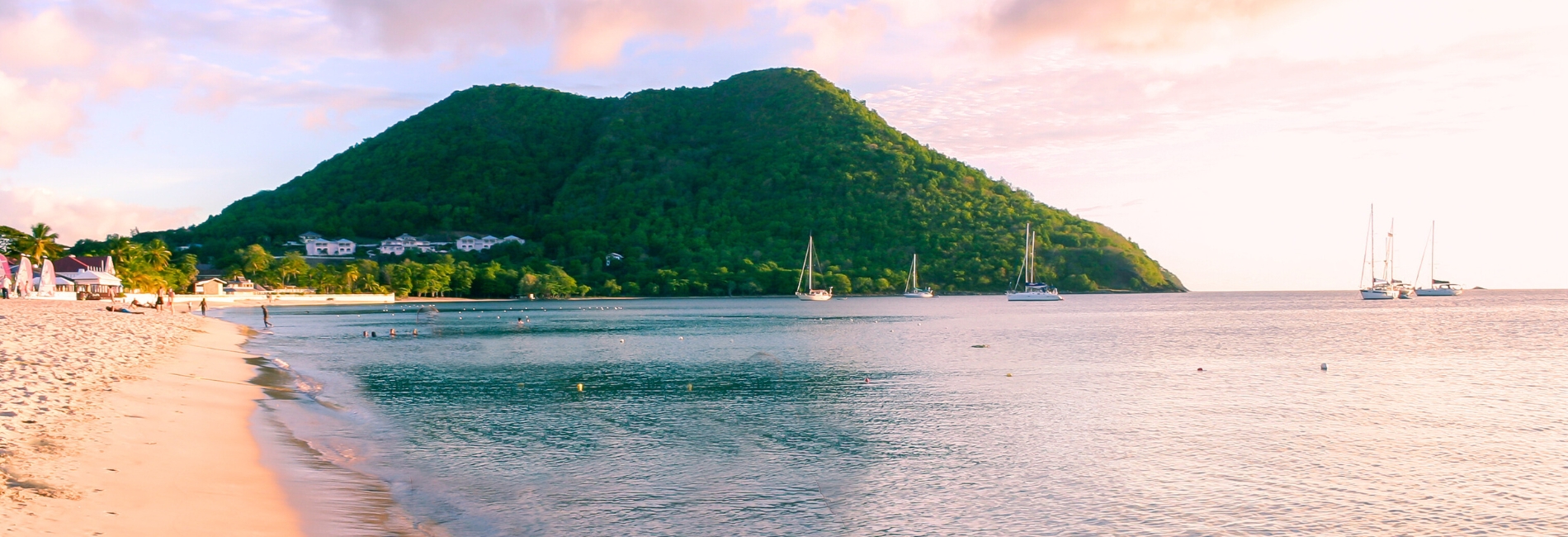 Beautiful beach in St. Lucia at sunset with white sand, calm turquoise water, sailboats anchored in bay, lush green mountains, palm trees, and pink sky