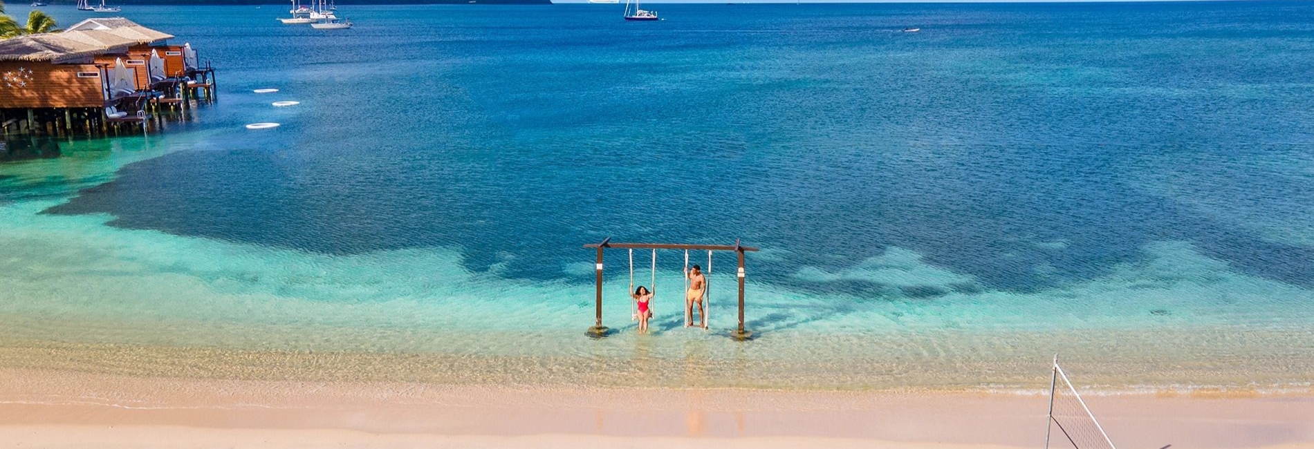 Stunning St. Lucia beach with crystal clear turquoise water, beach swings in shallow water, white sand beach, overwater bungalows, sailboats in distance, and volleyball net
