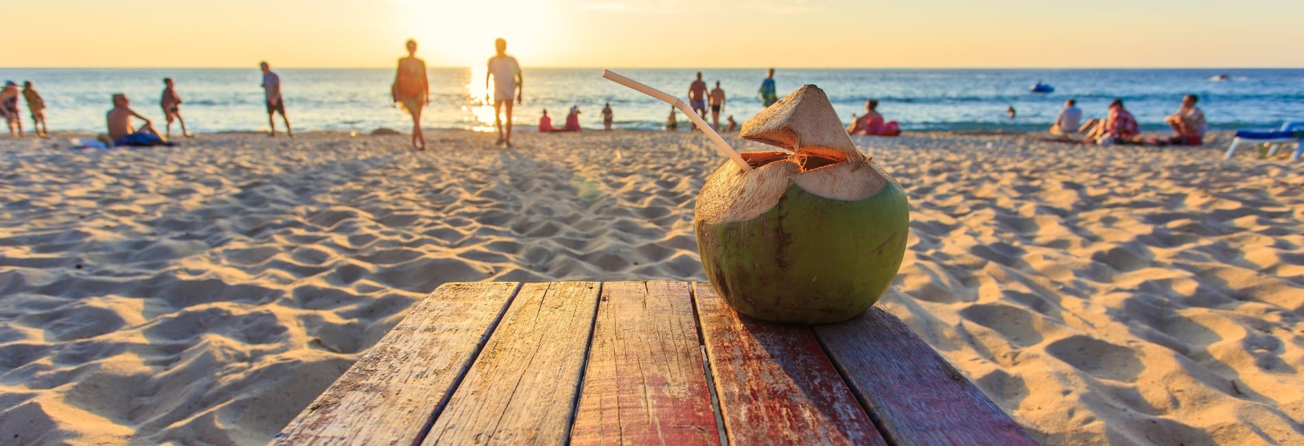 Fresh coconut drink with straw on wooden table at Reduit Beach in St. Lucia with golden sunset and beachgoers enjoying the sand and Caribbean Sea in background